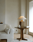 Old wood table lamp with linen shade on wooden table in cozy living room with soft beige couch and large window.