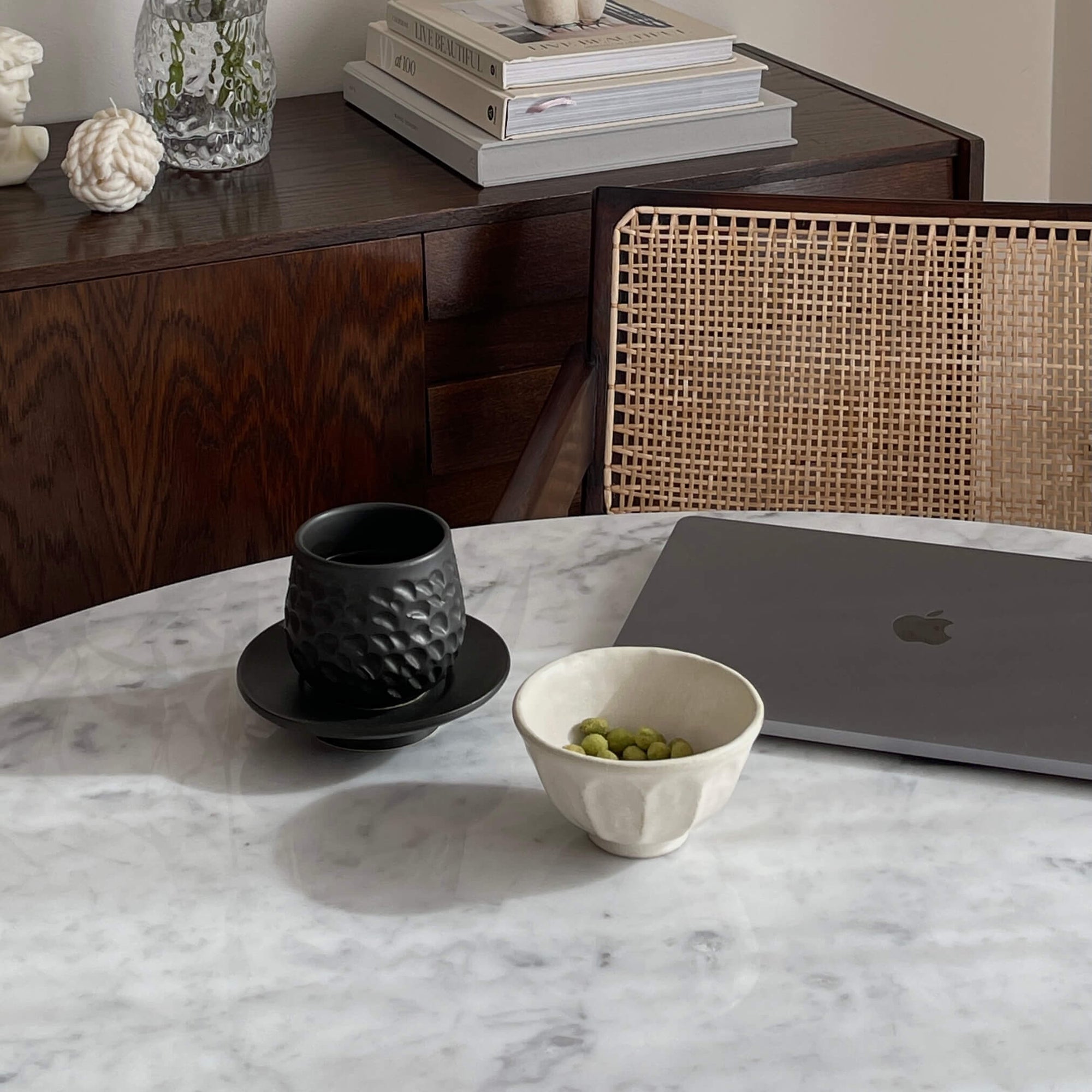Marble table with a black cup and saucer, white bowl of peas, and a laptop, against a woven chair and wooden sideboard setup.