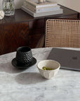 Marble table with a black cup and saucer, white bowl of peas, and a laptop, against a woven chair and wooden sideboard setup.