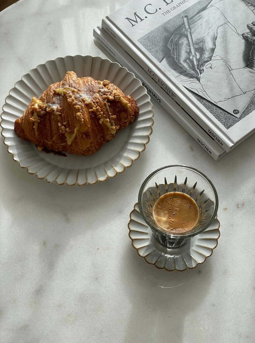 Elegant porcelain plate with croissant and a cup of espresso on a marble table next to art books.