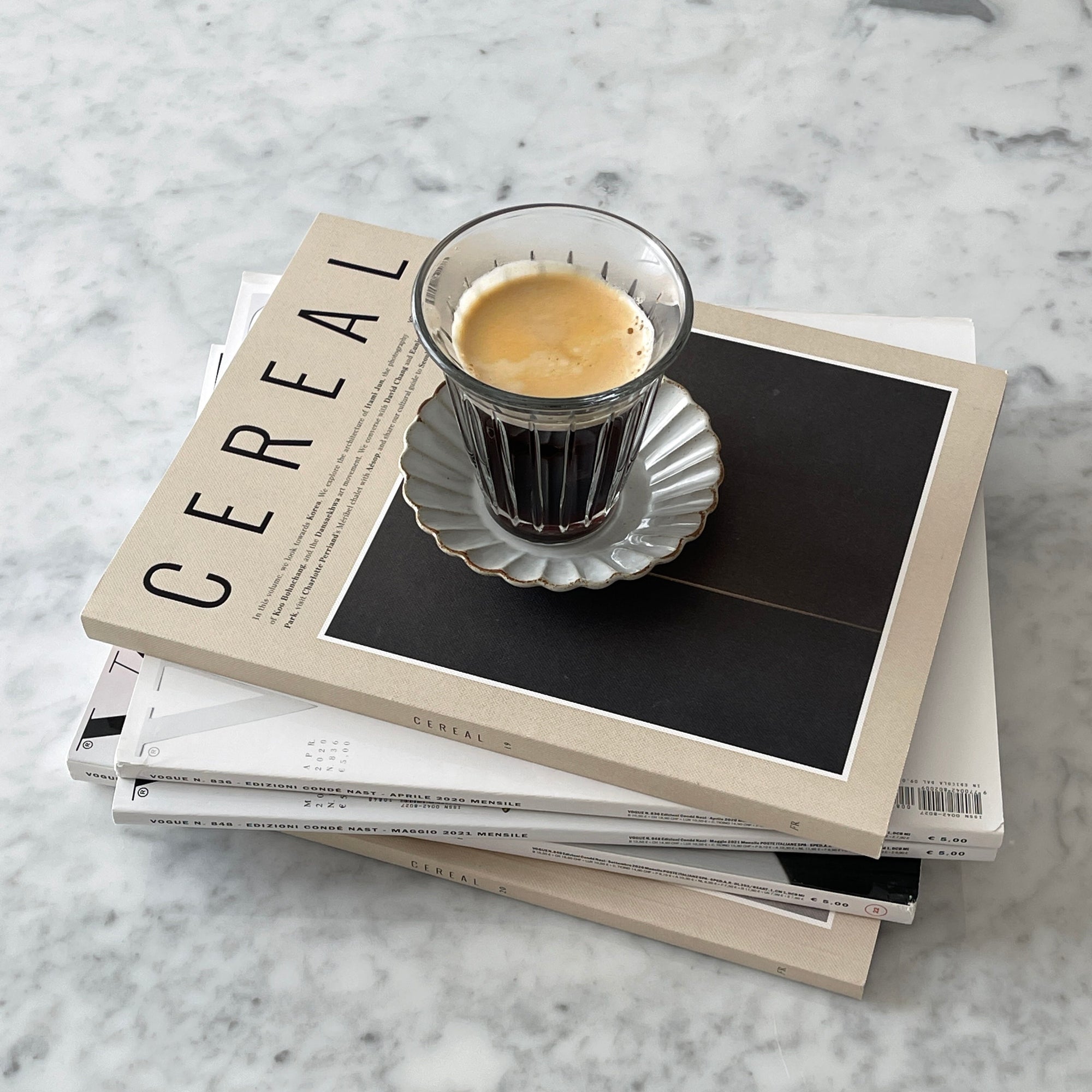 Zinc coffee glass cup with espresso on stack of Cereal magazines on marble countertop, French brasserie inspired design.