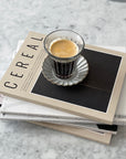 Zinc coffee glass cup with espresso on stack of Cereal magazines on marble countertop, French brasserie inspired design.