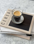 Zinc coffee glass cup with espresso on stack of Cereal magazines on marble countertop, French brasserie inspired design.