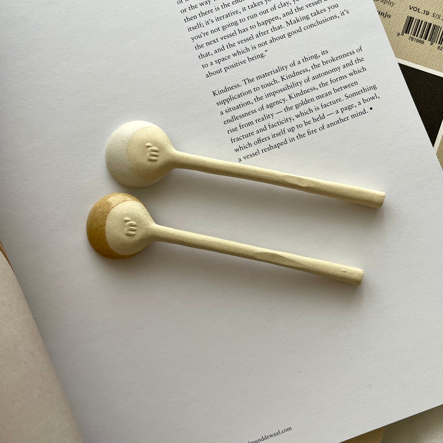 Two Marumitsu Bean Spoons with semi-matte rustic glaze tips on a book, showcasing a unique bean-shaped design and white clay finish.