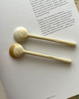 Two Marumitsu Bean Spoons with semi-matte rustic glaze tips on a book, showcasing a unique bean-shaped design and white clay finish.