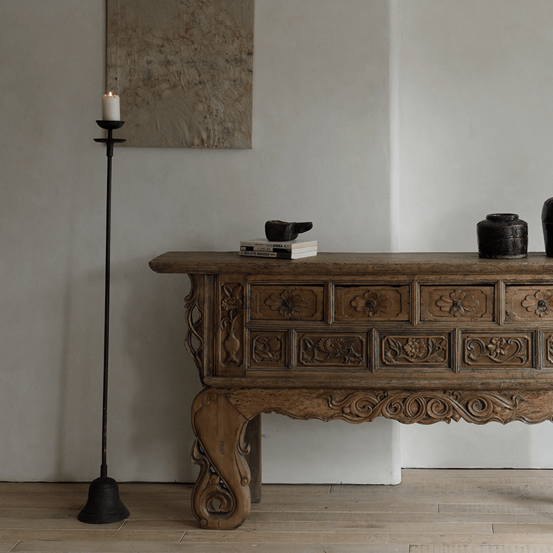 Aimu floor candle holder with antique finish beside an ornate wooden console table in a stylish, rustic living room setting.