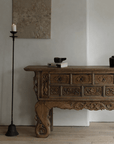 Aimu floor candle holder with antique finish beside an ornate wooden console table in a stylish, rustic living room setting.