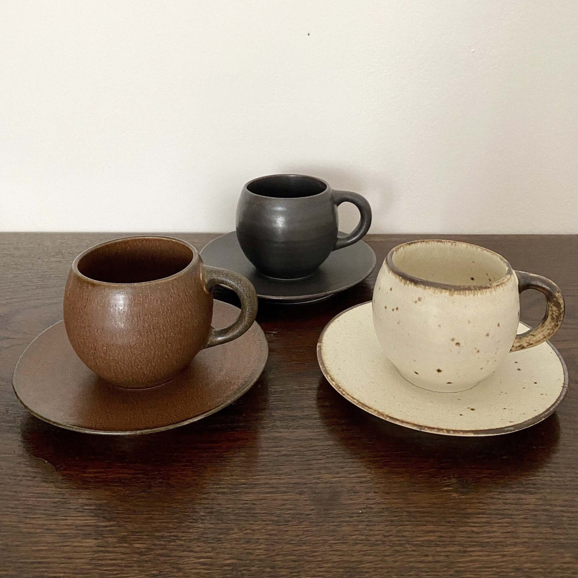 Three textured porcelain mugs with matching saucers in brown, black, and ivory displayed on a wooden surface.