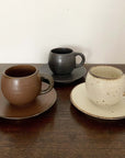 Three textured porcelain mugs with matching saucers in brown, black, and ivory displayed on a wooden surface.