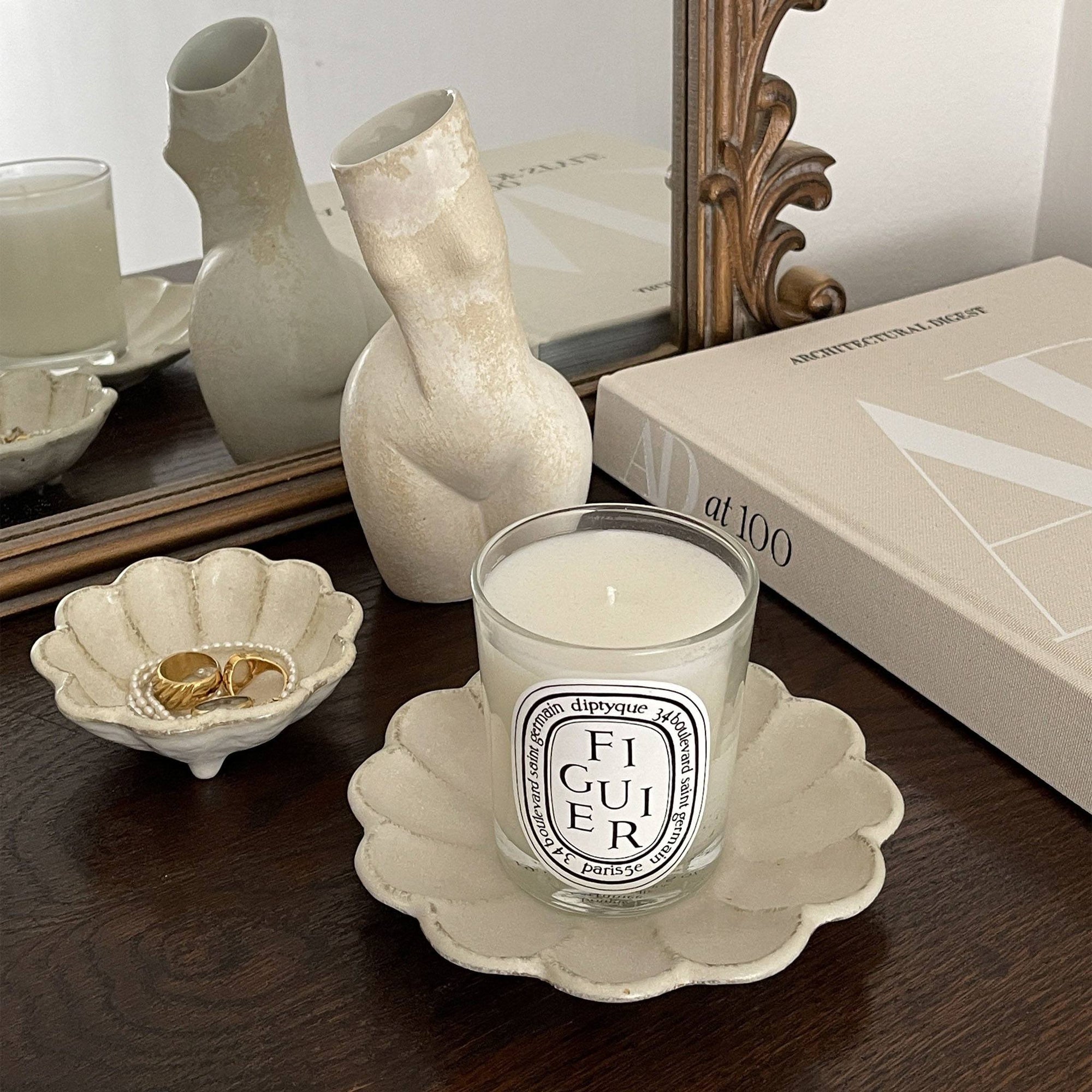 Elegant beige ceramic candle holder with scented candle beside decorative vase and book on wooden table.