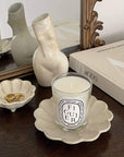 Elegant beige ceramic candle holder with scented candle beside decorative vase and book on wooden table.