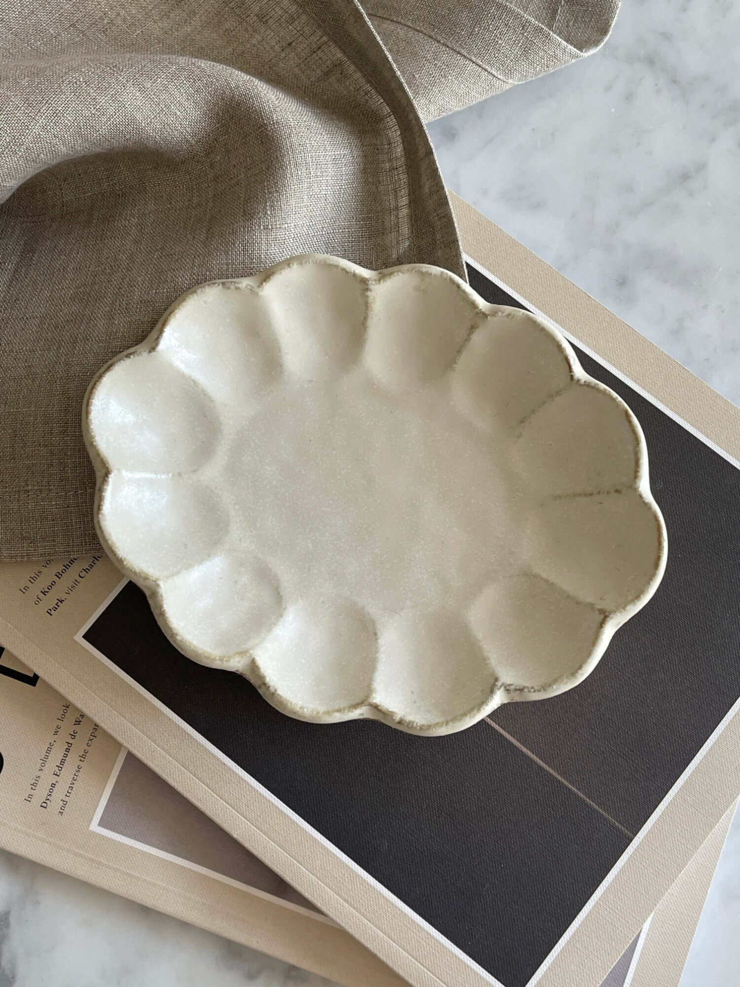 Porcelain Rinka Oval Plate with flower shape from Kaneko Kohyo, set on a stack of magazines, showcasing its delicate craftsmanship.