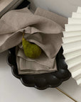 Black bowl with scalloped edges holding beige linen napkins and a green pear, set on a white surface near a white paper fan.