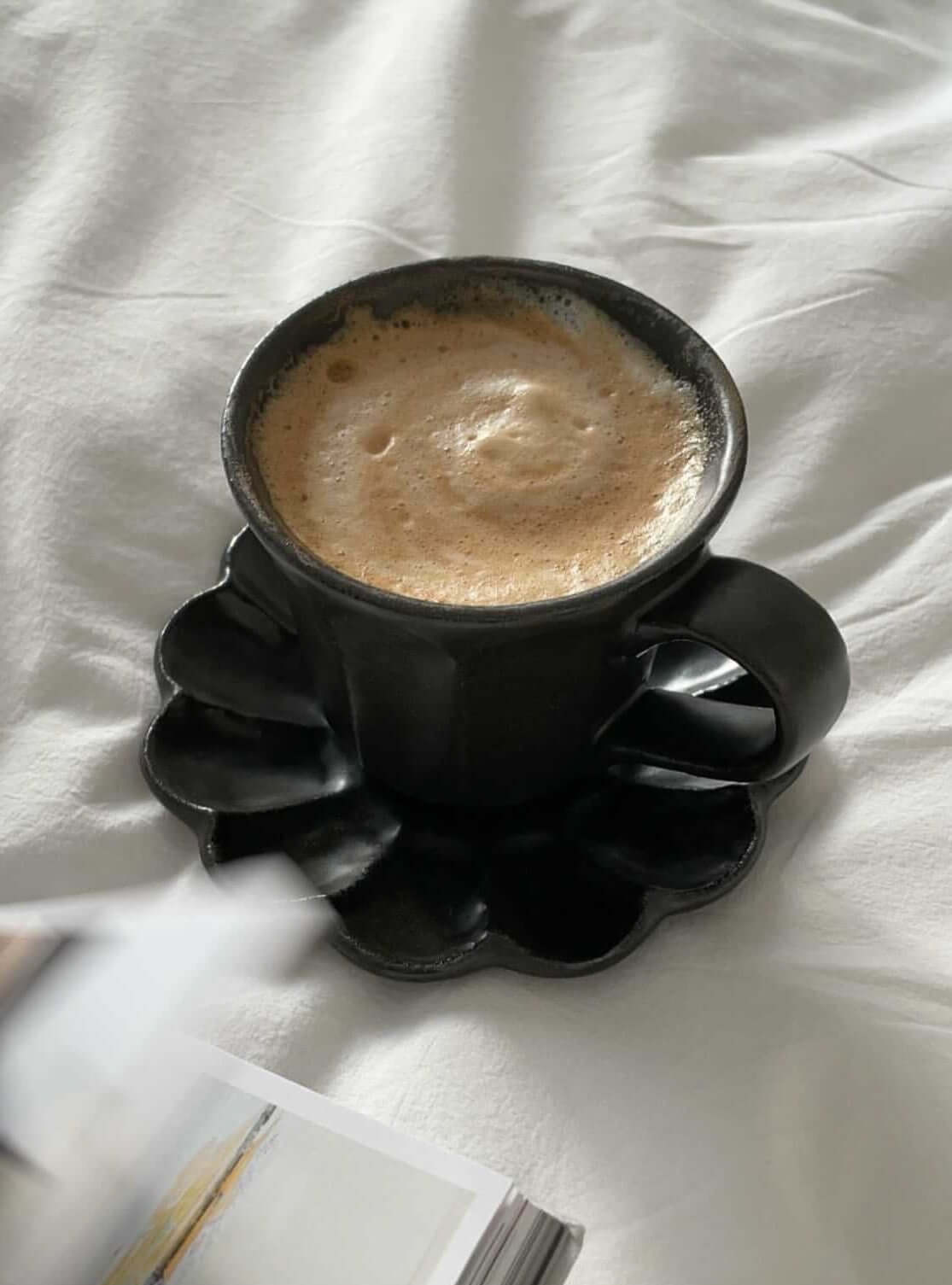 Black Rinka mug with frothy coffee on matching scalloped plate, set on a white textured surface. Elegant handmade Japanese porcelain.
