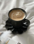 Black Rinka mug with frothy coffee on matching scalloped plate, set on a white textured surface. Elegant handmade Japanese porcelain.