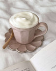 Rinka Mug by Kaneko Kohyo with frothy drink on a flower-shaped saucer, next to an open book on a white background.