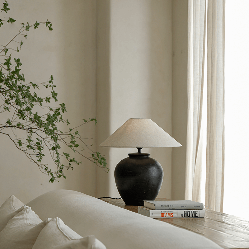 Elegant egg-shaped clay pot table lamp with linen shade on a wooden table, next to stacked books and greenery in a serene room setting.