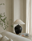 Elegant egg-shaped clay pot table lamp with linen shade on a wooden table, next to stacked books and greenery in a serene room setting.