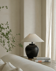 Elegant egg-shaped clay pot table lamp with linen shade on a wooden table, next to stacked books and greenery in a serene room setting.