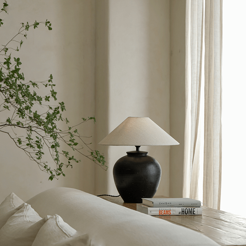 Egg-shaped clay pot table lamp with linen shade on wooden surface near books and sofa, creating a serene and cozy atmosphere.