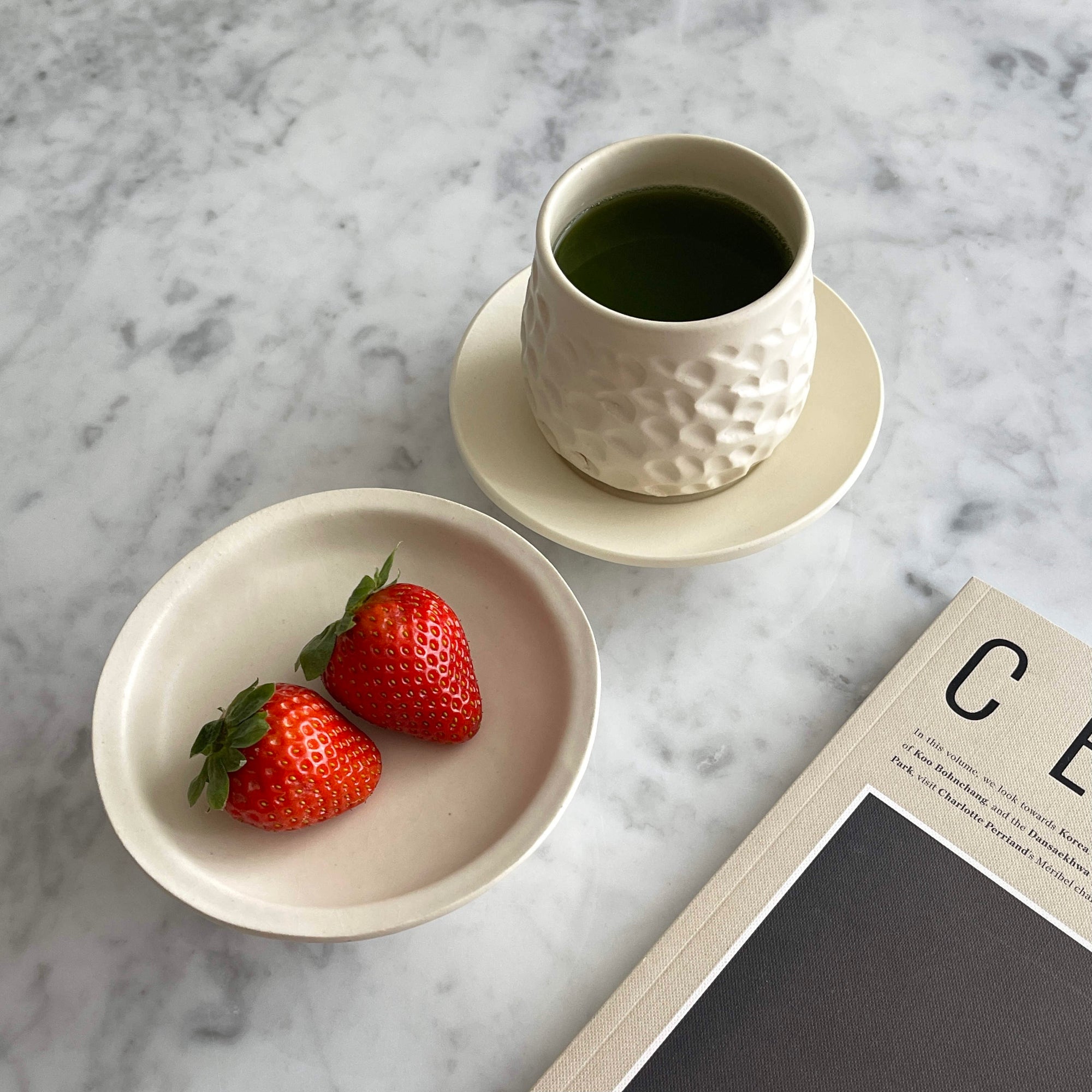 Ceramic cup with green tea, white plate with strawberries, and magazine on marble table.