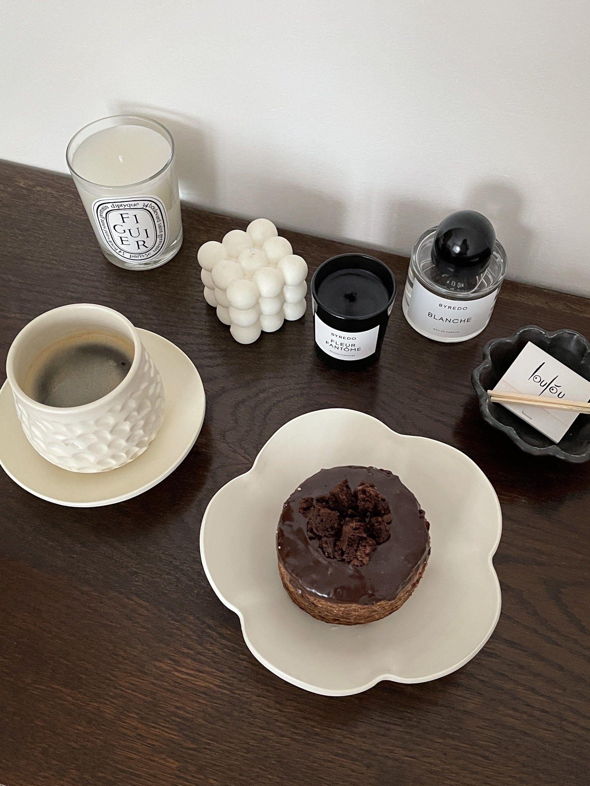 Elegant table setting with a coffee cup and a chocolate pastry on a unique flower-shaped plate, surrounded by candles and decor.