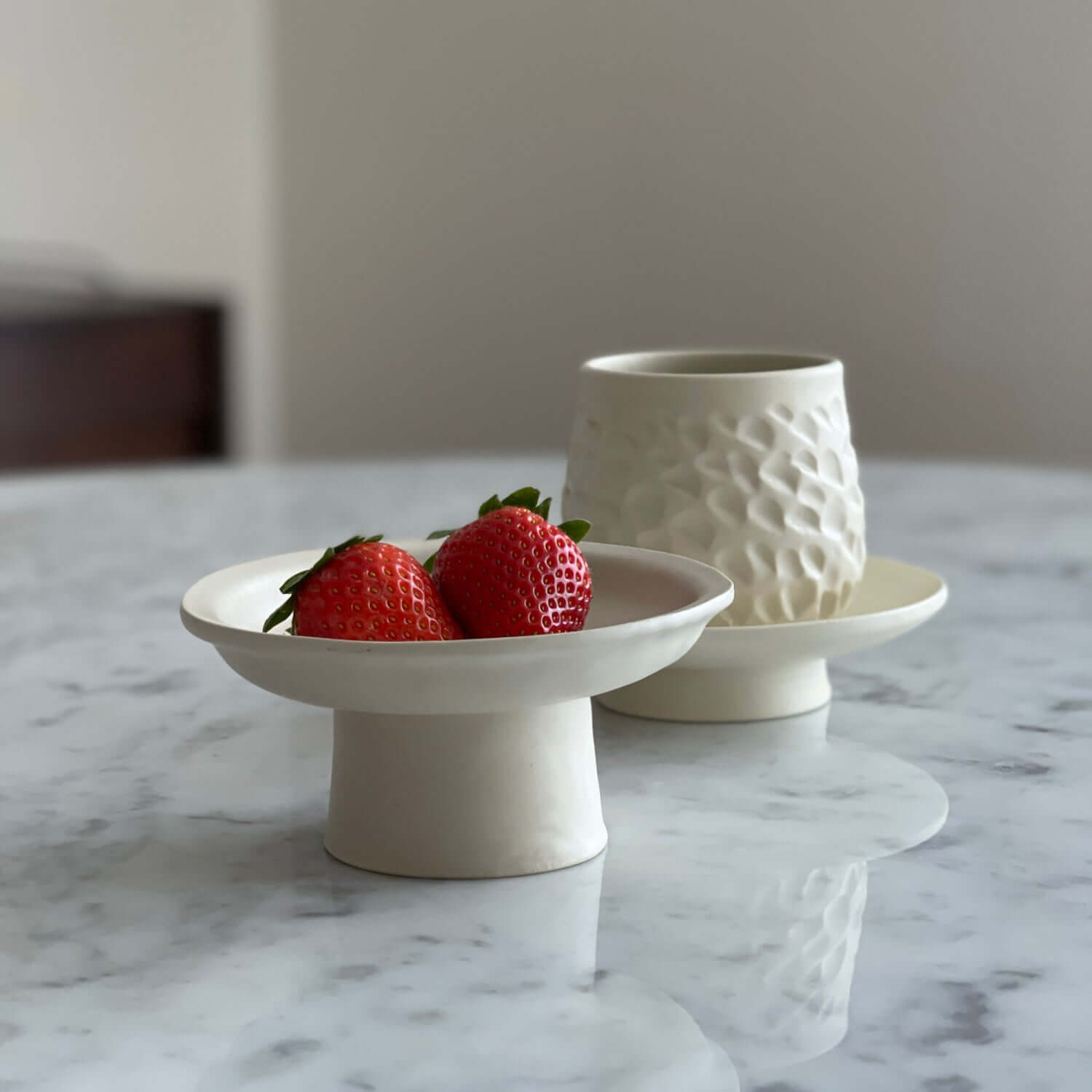 Ceramic pedestal dish with strawberries and a textured ceramic cup on a marble table.