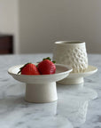 Ceramic pedestal dish with strawberries and a textured ceramic cup on a marble table.