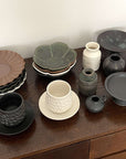 Shinogi Cup and Saucer set on a wooden table with assorted pottery plates and vases, showcasing elegant, nature-inspired designs.