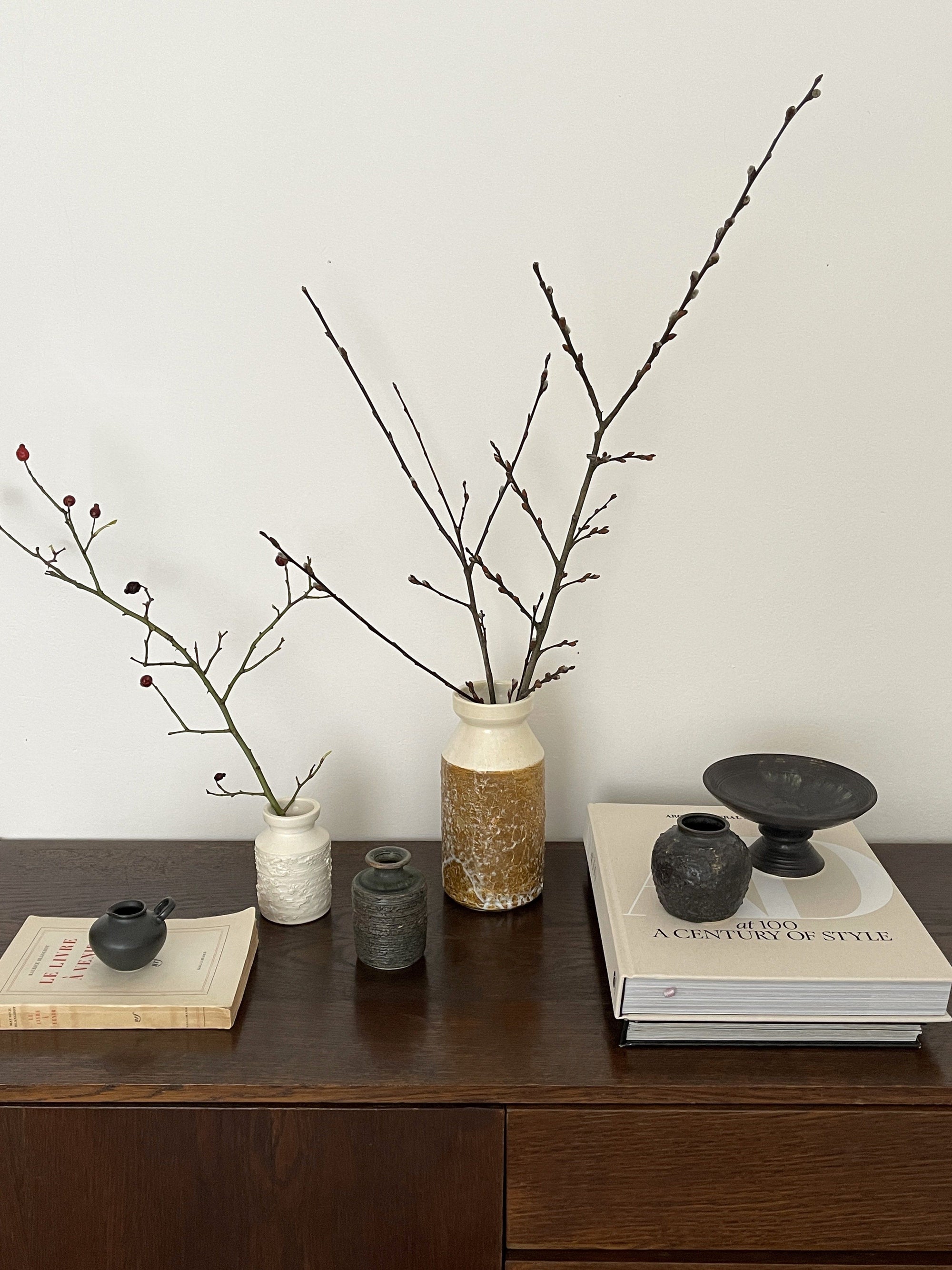 Ceramic vases with branches on a wooden table, accompanied by books and pottery, creating a minimalist and natural decor setting.