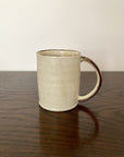 Rila Ivory Straight Mug, textured porcelain, microwave-safe, stackable, warm matte finish, compact size, on dark wood surface.