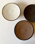 Trio of textured Kaneko Kohei porcelain plates in ivory, brown, and black, showcasing warm, slightly rough finishes, 11.5cm diameter.