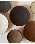 Assorted textured porcelain plates in neutral tones, featuring ivory, black, and brown hues, showcasing Kaneko Kohyo craftsmanship.