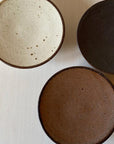 Three textured porcelain plates in cream, black, and brown by Kaneko Kohyo, showcasing matte finishes and rustic designs.