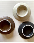 Three textured porcelain cups by Kaneko Kohei in ivory, brown, and black colors placed on a white surface.