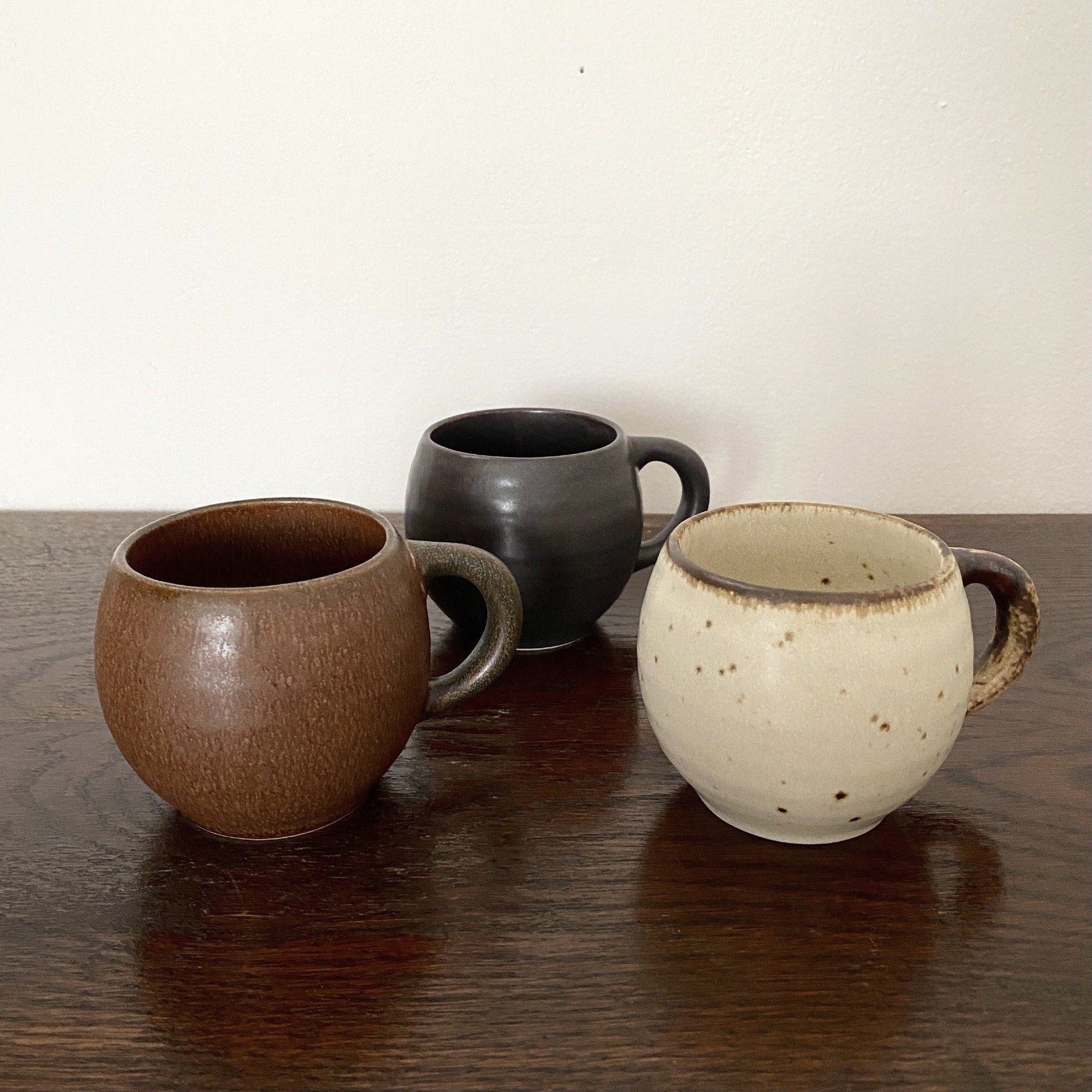 Three textured porcelain mugs in brown, black, and cream on a wooden surface, showcasing a rustic design and perfect palm fit.