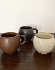 Set of three textured porcelain mugs in brown, black, and cream on wooden surface.