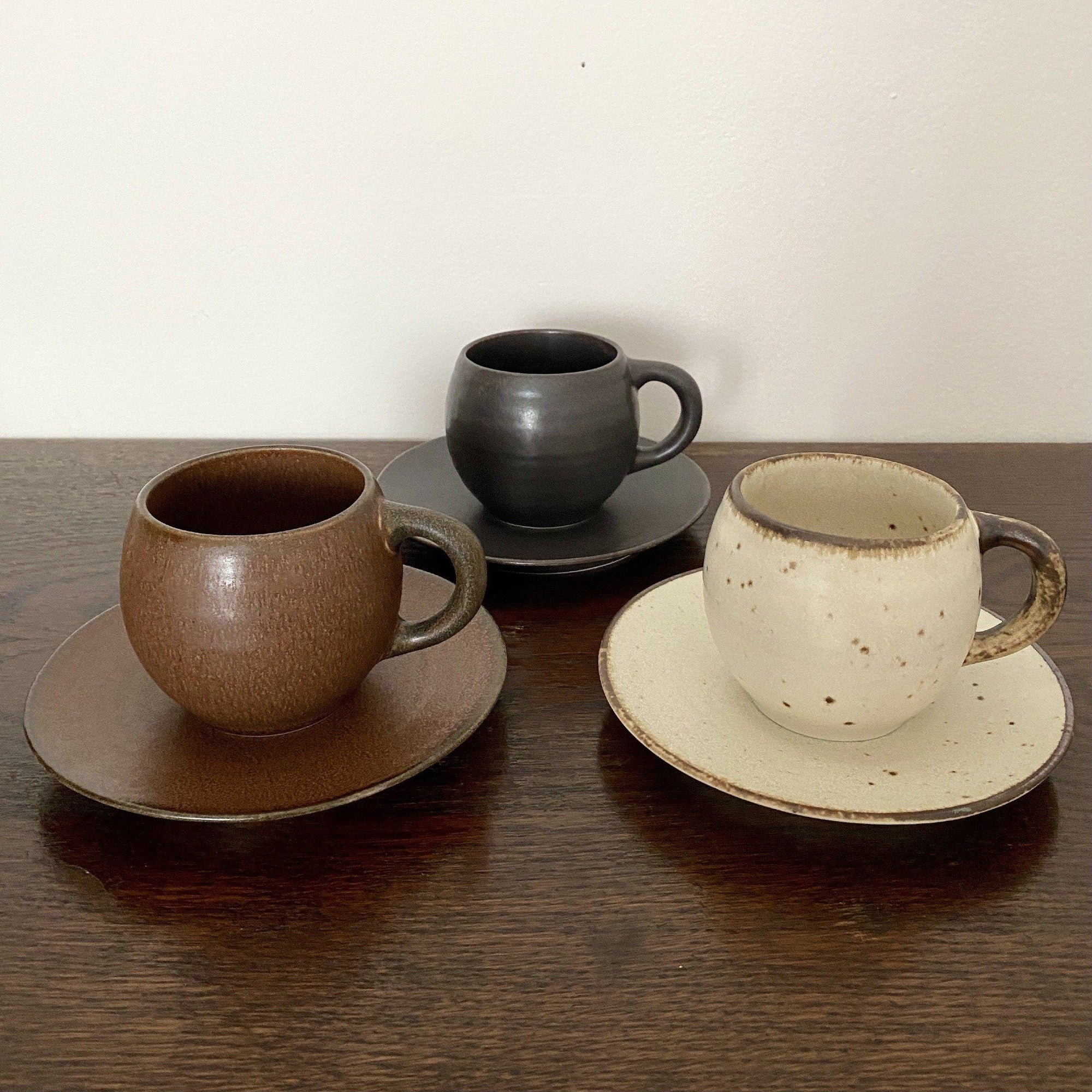 Three textured porcelain mugs and saucers in brown, black, and cream, displayed on a wooden surface.