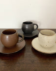 Three textured porcelain mugs and saucers in brown, black, and cream, displayed on a wooden surface.