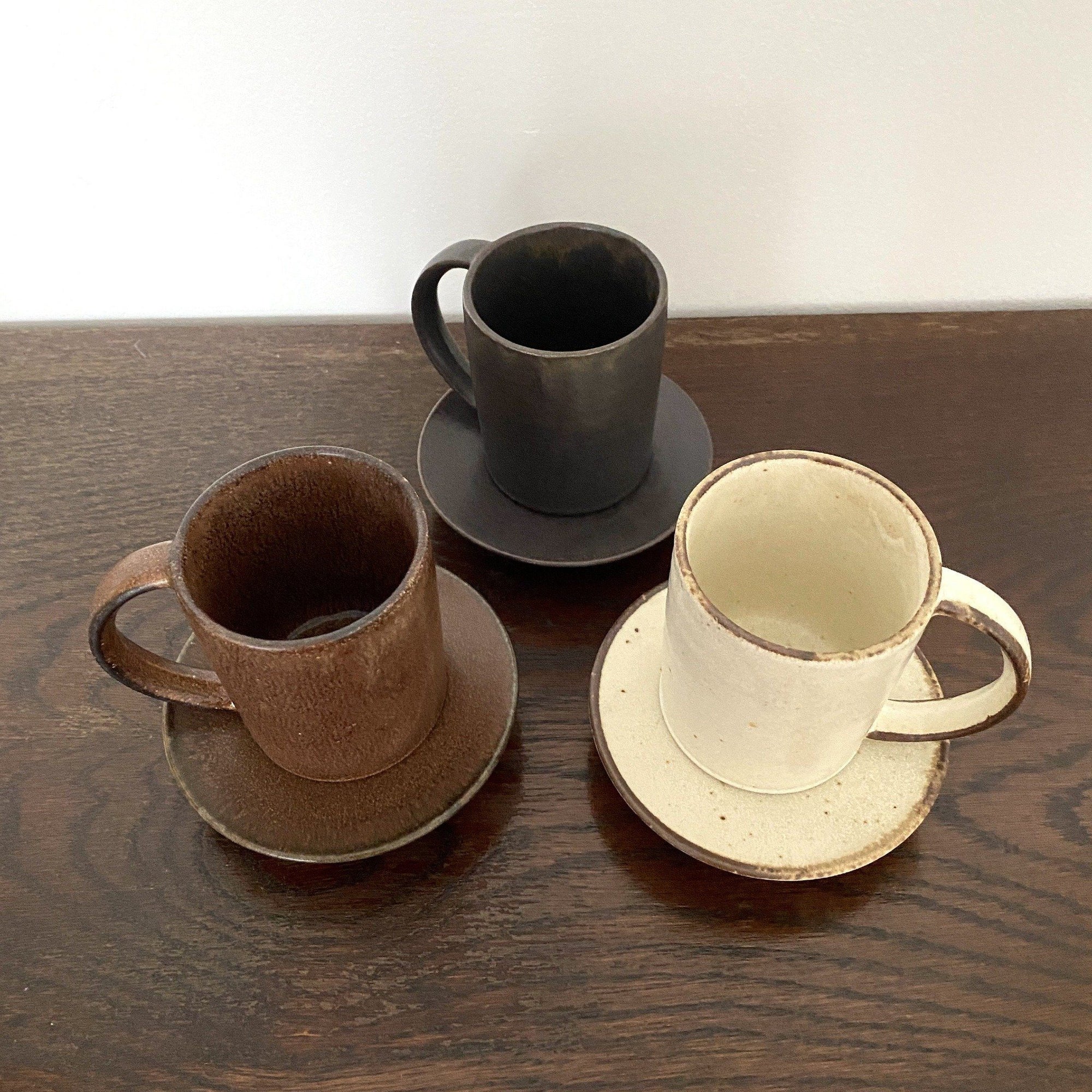 Three porcelain straight mugs in black, brown, and white on matching saucers from Kaneko Kohyo, displayed on a wooden surface.