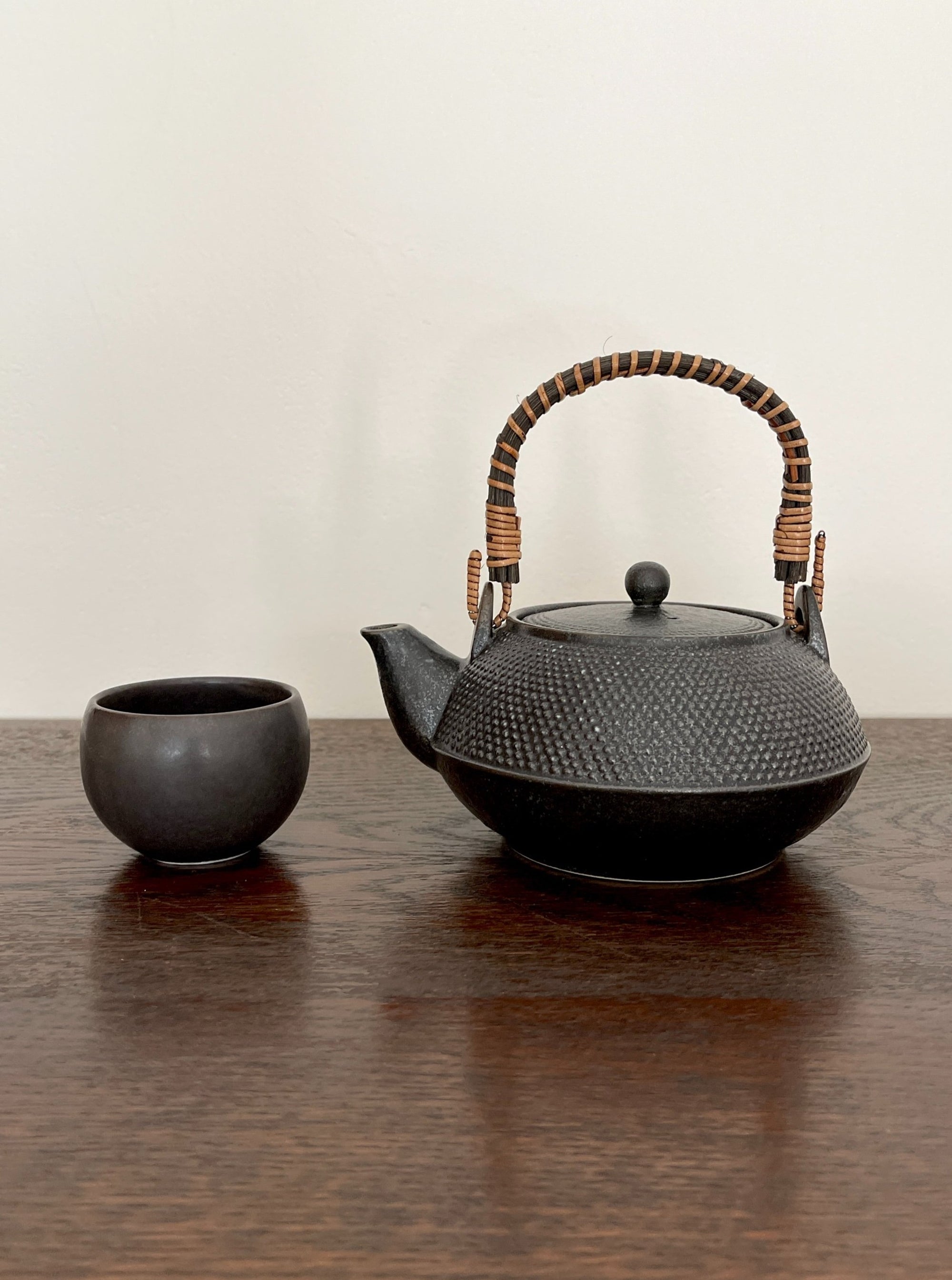 Black textured teapot with wicker handle next to a small black cup on a wooden table.