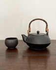 Black textured teapot with wicker handle next to a small black cup on a wooden table.