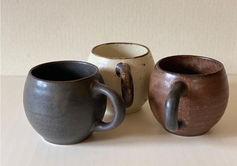 Three textured porcelain mugs in black, ivory, and brown displayed on a table, showcasing their distinct matte finishes and round handles.