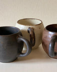 Three porcelain mugs from Kaneko Kohyo in dark brown, beige, and brown, showcasing round design and matte texture on a neutral background.