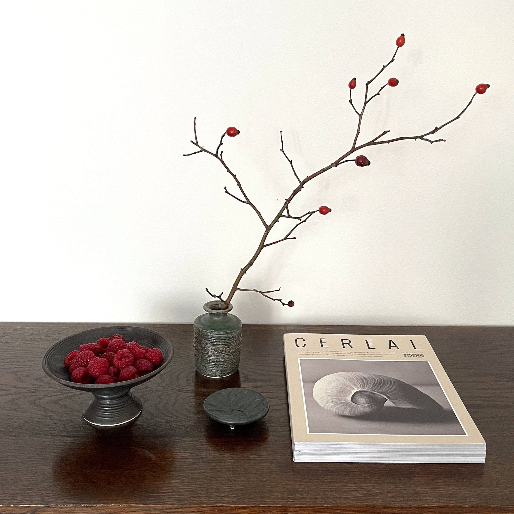 Elegant table setting with a branch in a green vase, a bowl of raspberries, and a chic magazine, embodying sophisticated home decor.