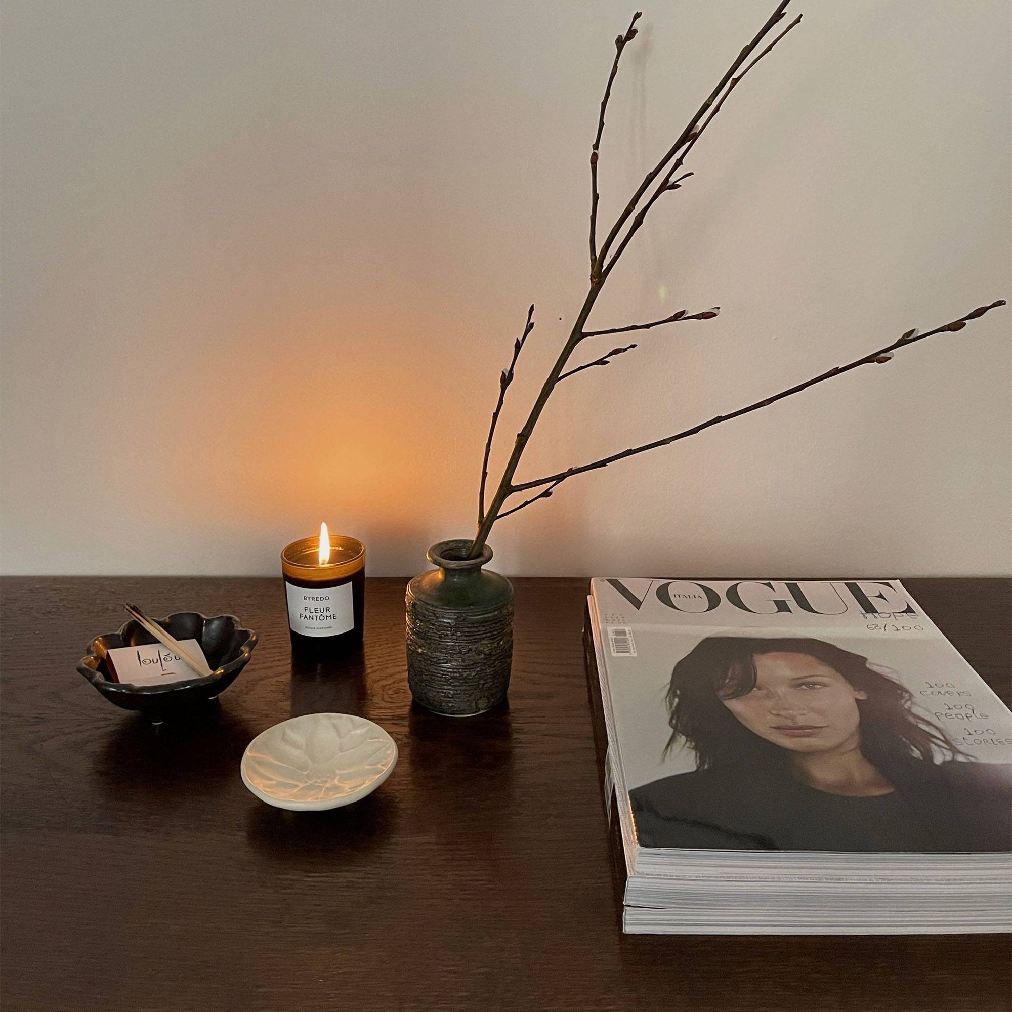 Decorative table setting with a green vase, candle, twigs, and magazines on a wooden surface, creating a cozy ambiance.
