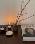 Decorative table setting with a green vase, candle, twigs, and magazines on a wooden surface, creating a cozy ambiance.