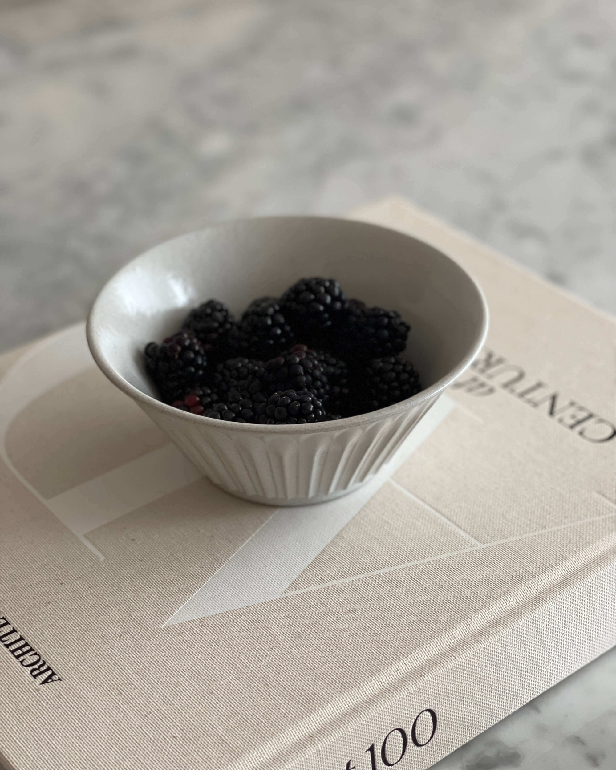 White porcelain bowl filled with blackberries set on a book on a marble surface.