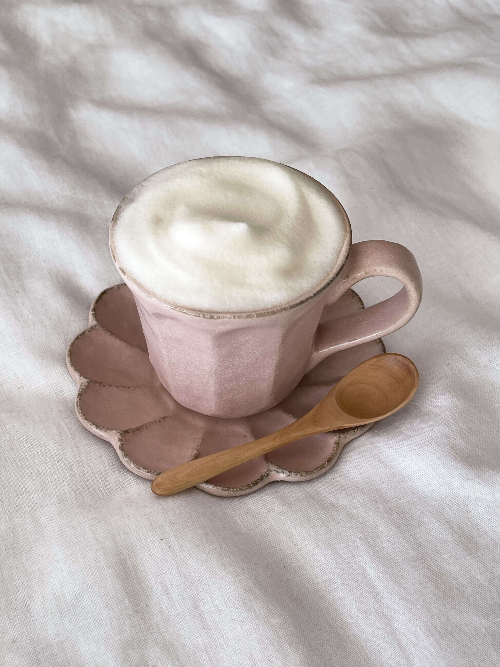 Pink ceramic cup with frothy coffee on scalloped saucer, accompanied by wooden teaspoon.