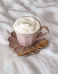 Pink ceramic cup with frothy coffee on scalloped saucer, accompanied by wooden teaspoon.